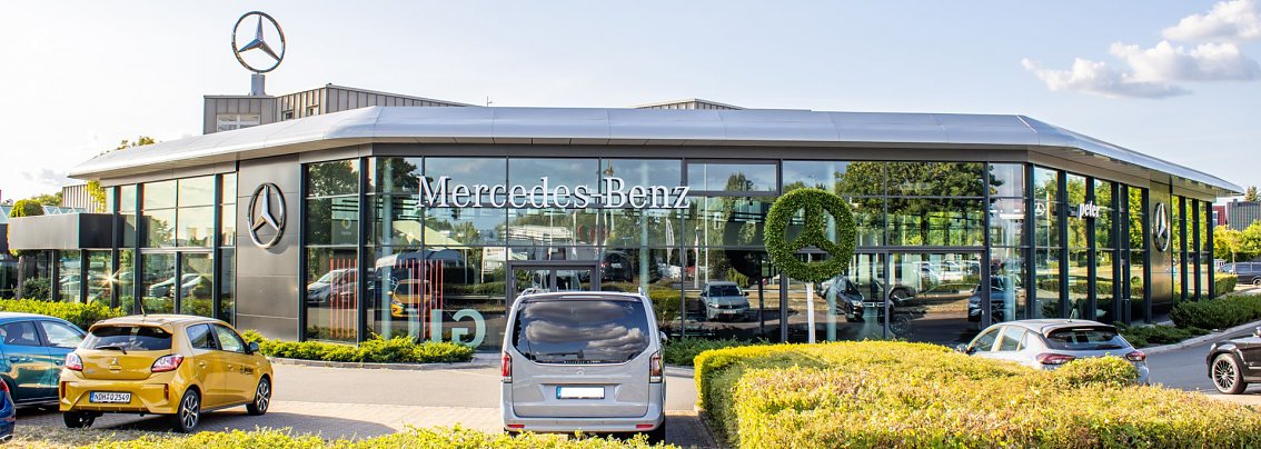 Au&szlig;enansicht eines modernen Mercedes-Benz Autohauses Peter in Nordhausen mit gro&szlig;fl&auml;chiger Glasfassade, Mercedes-Benz Logos auf dem Dach und als Heckenschnitt, mehrere schwarze Fahrzeuge im Vordergrund, bew&ouml;lkter Himmel.