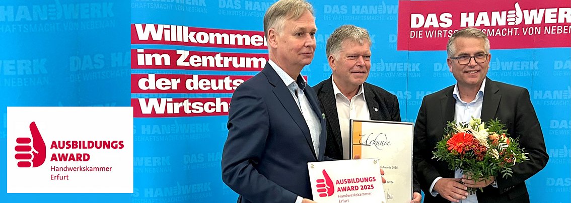 Drei Männer stehen vor einer blauen Wand mit dem Schriftzug „Das Handwerk  Die Wirtschaftsmacht von nebenan“. Einer hält eine Urkunde, ein anderer eine Blumenstrauß, der dritte das Schild „Ausbildungsaward 2025 Handwerkskammer Erfurt“. Das Bild zeigt die Übergabe des Ausbildungsawards an die Autohaus Peter Gruppe.