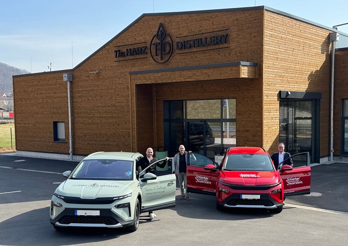 Zwei vollelektrische SUV mit Markenbeklebung � ein gr&uuml;ner f&uuml;r The HARZ DISTILLERY und ein roter f&uuml;r Schierker Feuerstein � stehen vor einem modernen Holzgeb&auml;ude. Drei M&auml;nner pr&auml;sentieren die Fahrzeuge mit ge&ouml;ffneten T&uuml;ren.