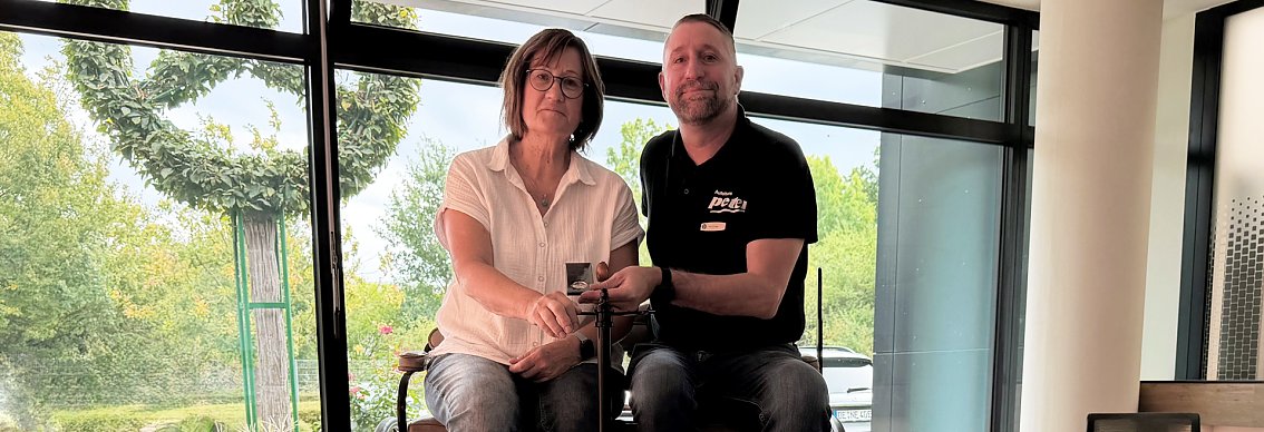 Eine Kundin bekommt vom Mitarbeiter des Autohauses den exklusiven goldenen Mercedes-Benz-Schl&uuml;sselanh&auml;nger &uuml;berreicht. Beide sitzen auf dem Patent-Motorwagen, den man aber nicht vollst&auml;ndig sieht.