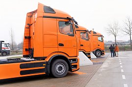 Seitenansicht von drei orangefarbenen Mercedes-Benz eActros 600 Lkw, die in einer Reihe auf einem Hof stehen. Rechts im Bild sind drei Personen im Gespräch. (Foto: ex animo MEDIA)