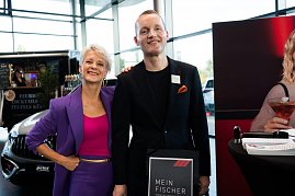 Eine Frau (Moderatorin Mandy Kossira) in lila Blazer und pinkem Top steht neben einem Mann im schwarzen Anzug. Beide lächeln. Neben ihnen ein Aufsteller mit der Aufschrift Mein Fischer.