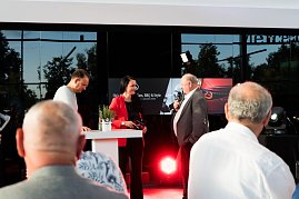 Christina Dost im roten Blazer spricht mit Helmut Peter im grauen Anzug. Beide stehen auf einer Bühne im Autohaus, links daneben, ebenfalls auf der Bühne, Andreas Peter. Das Banner He’s Mercedes – Cars, BBQ & Style ist im Hintergrund zu sehen.