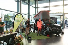 Ein schwarzer Mercedes-Camper mit geöffnetem Aufstelldach steht auf grünem Kunstrasen im Autohaus. Daneben zwei Männer, einer zeigt den Daumen hoch. Links im Bild ein Verkaufsstand mit Prospekten und Produkten.