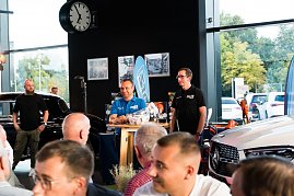 Zwei Männer stehen an einem kleinen Tisch, einer trägt ein blaues Hemd mit DTZ-Logo, der andere ein schwarzes Polohemd. Sie präsentieren Produkte, im Hintergrund sind ein schwarzer Mercedes und eine große Uhr zu sehen.