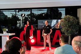 Christina Dost im roten Blazer spricht mit einem Mikrofon auf der Bühne. Neben ihr steht ein Mann im schwarzen Polohemd mit Autohaus-Logo. Hinter ihnen eine große LED-Wand mit He’s Mercedes – Cars, BBQ & Style.
