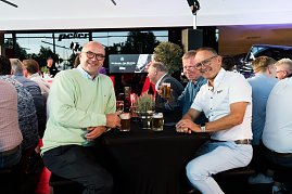 Drei Männer sitzen an einem Stehtisch mit Getränken und lachen in die Kamera. Viele weitere Gäste sitzen im Hintergrund an Tischen.