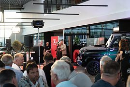 Blick in die Eventhalle mit vielen Gästen im Vordergrund. Auf der Bühne stehen Christina Dost im roten Blazer und ein Mann in beiger Jacke, daneben ein schwarzer Mercedes-Geländewagen.