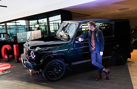 Ein junger Mann lehnt entspannt an einem schwarzen Mercedes-Geländewagen und schaut zur Seite. Der Wagen ist im Ausstellungsbereich mit Scheinwerfern beleuchtet.