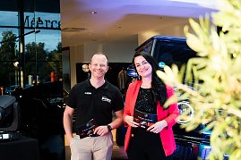 Eine Frau im roten Blazer (Christina Dost) und ein Mann im schwarzen Polohemd (Carsten Dost) stehen nebeneinander und lächeln. Beide halten Moderationskarten, im Hintergrund ein Mercedes-Geländewagen.