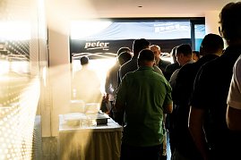 Eine Gruppe Männer steht in einer Schlange am Buffet. Sonnenlicht fällt durch die Tür, im Hintergrund ein Pavillon mit Autohaus-Peter-Logo.