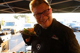 Ein Koch mit Brille und schwarzer Schürze mit Mercedes-Logo hält lächelnd ein belegtes Sandwich in die Kamera. Im Hintergrund ein Foodtruck und Fahrzeuge im Sonnenlicht.