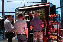 Mehrere Männer stehen an einem Gin-Stand mit schwarzem Verkaufswagen. Auf Schildern steht Luzifer Gin und Gin-Cocktails. Regale mit Flaschen sind daneben aufgestellt.