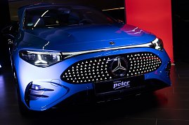 Nahaufnahme der Front eines blauen Mercedes CLA mit beleuchtetem Kühlergrill im Showroom. Ein rotes Eventbanner mit Mercedes-Logo ist im Hintergrund zu sehen.