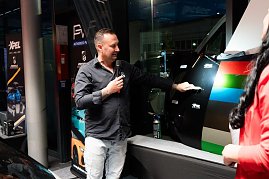 Ein Mann in schwarzem Hemd erklärt mit Mikrofon verschiedene Lack- und Folienmuster an einer Autotür. Daneben bunte Farbfolienstreifen.