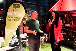 Christina Dost im roten Blazer moderiert neben einem Mann im grünen T-Shirt vor einem gelben Banner mit der Aufschrift Möbius – Grün macht glücklich!. Hinter ihnen ein schwarzer Mercedes-Camper.