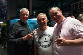 Drei Männer stehen nebeneinander und lachen in die Kamera. Einer trägt ein weißes Mercedes-Benz-T-Shirt, die anderen dunkle Hemden. Alle haben Getränke in der Hand.