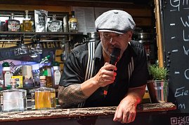 Ein Mann mit Schiebermütze und Tattoos spricht ins Mikrofon an einem Gin-Stand. Vor ihm mehrere Flaschen verschiedener Spirituosen.