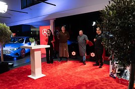 Christina Dost roten Blazer moderiert auf der Bühne. Neben ihr stehen drei Köche und Grillmeister in Schürzen und Arbeitskleidung. Im Hintergrund ein blauer Mercedes.