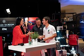 Christina Dost im roten Blazer interviewt einen jungen Mann im weißen Mercedes-AMG-Petronas-Shirt. Beide lachen. Dahinter ein Stand mit Mercedes-Merchandise.