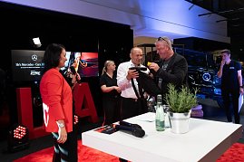 Ein Mann in weißem Hemd und ein weiterer in schwarzer Kleidung packen gemeinsam ein Geschenk aus. Links steht Christina Dost im roten Blazer mit Mikrofon.