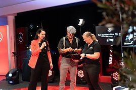 Christina Dost steht mit einem Mann in Schiebermütze und einer Frau im schwarzen Polohemd auf der Bühne. Gemeinsam ziehen sie Lose aus einer Schale.