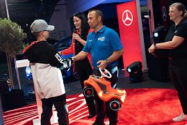 Ein Junge mit Mercedes-Cap bekommt ein rotes Bobbycar von einem Mann im blauen DTZ-Shirt überreicht.