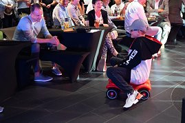 Ein Junge im schwarzen AMG-Hoodie und Mercedes-Cap fährt lachend auf einem roten Bobbycar durch den Showroom. Erwachsene Gäste sitzen an Tischen und beobachten ihn amüsiert.