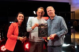 Christina Dost im roten Blazer steht mit Andreas Peter und einem Gewinner auf der Bühne. Gemeinsam halten sie einen großen Gutschein mit Nürburgring-Aufdruck in die Kamera.