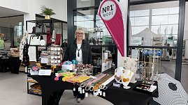Stand von No1 Mode im Autohaus Autowelt Peter. Eine Frau präsentiert Taschen, Tücher, Schmuck und Accessoires, die auf Tischen und Regalen dekoriert sind. Daneben steht eine pinkfarbene Werbefahne mit dem Logo des Modegeschäfts.