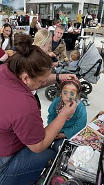 Kinderschminken im Autohaus Autowelt Peter: Ein Mädchen mit Katzenohren-Stirnband bekommt ein buntes Regenbogen-Gesicht gemalt. Im Hintergrund sitzen weitere Familien mit Kindern.