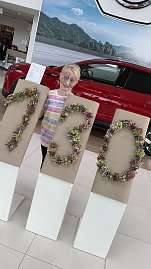 Ein Kind mit bunt bemaltem Gesicht steht im Autohaus Autowelt Peter hinter großen Ziffern aus Blumen, die ‚130‘ formen – passend zum Jubiläum 130 Jahre Škoda. Daneben ist ein rotes Auto ausgestellt.