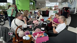 Frauen sitzen an einem Tisch im Autohaus Autowelt Peter und stricken gemeinsam. Auf dem Tisch liegen bunte Wollknäuel, Strickutensilien und Handarbeiten. Im Hintergrund sind weitere Gäste und Ausstellungsstände zu sehen.