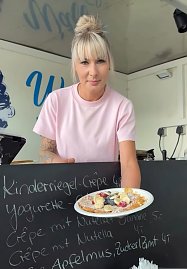 Frau in rosa T-Shirt präsentiert einen frisch zubereiteten Crêpe mit Obst und Puderzucker. Im Hintergrund ist eine Tafel mit Crêpe-Sorten beschrieben.