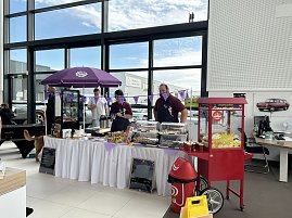 Verkaufsstand mit Snacks und Süßwaren im Autohaus Autowelt Peter. Unter einem lila Sonnenschirm stehen Verkäufer, daneben eine rote Popcornmaschine.