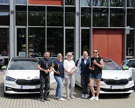 Fünf Personen stehen vor einem modernen Gebäude und posieren mit Champagnerflaschen vor zwei weißen koda-Fahrzeugen. Auf den Autos sind grüne Elemente und das Logo der „Praxisklinik für Zahnheilkunde“ zu sehen.