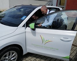 Ein Mann im blauen Sakko zeigt einer Frau auf dem Fahrersitz eines weißen koda-Fahrzeugs etwas auf einem Tablet. Auf der Autotür ist das Logo der „Praxisklinik für Zahnheilkunde“ zu sehen.