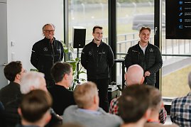 Drei Männer in schwarzen Jacken mit dem Mercedes-Benz 'Driving Experience'-Logo stehen vor einer Personengruppe und sprechen zu einem Publikum. Der Raum hat große Fenster, durch die man die Rennstrecke sieht.
