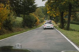 Eine Gruppe von Fahrzeugen fährt auf einer kurvigen Landstraße, die von Bäumen gesäumt ist. Im Vordergrund ist ein weißer Mercedes-Benz Cabrio zu sehen, gefolgt von weiteren Fahrzeugen. Die Bäume zeigen herbstliche Farben, und die Straße ist von grünen Wiesen und einem hügeligen Hintergrund umgeben.