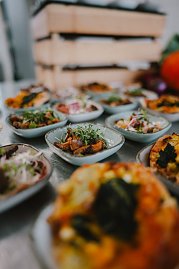 Mehrere kleine Schalen mit verschiedenen Gerichten sind auf einem Tisch arrangiert. Die Schalen enthalten bunte, frisch zubereitete Speisen, die mit Kräutern garniert sind. Im Hintergrund ist ein Stapel von Holzkisten sichtbar und einige weitere Speisen sind unscharf zu sehen.