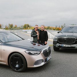 Zwei Männer stehen lächelnd nebeneinander zwischen zwei Mercedes-Maybach Fahrzeugen auf dem Gelände der Mercedes-Benz Driving Experience am Bilster Berg. Im Hintergrund ist die Rennstrecke mit herbstlicher Landschaft zu sehen.