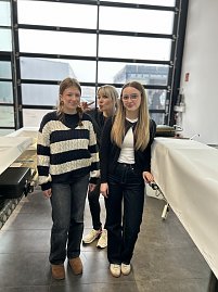 Drei junge Frauen stehen in einer Halle zwischen abgedeckten Tischen und schauen in die Kamera.