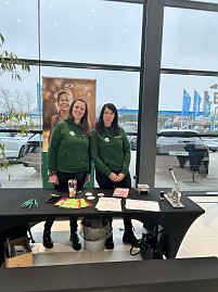 Zwei Frauen in gr&uuml;nen Hoodies stehen an einem Infotisch mit Stiften und Flyern, dahinter ein AOK-Plus-Banner. 