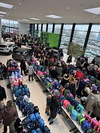 Blick von oben in ein volles Autohaus mit vielen Besuchern zwischen ausgestellten Autos und langen Tischen mit bunten Schulranzen.