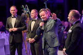 Redner mit Mikrofon vor elegant gekleideten G&auml;sten bei Gala-Event.