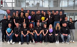 Man sieht ein Teamfoto mit vielen Menschen auf und vor einer kleinen Bühne. Alle haben das gleiche T-Shirt an, manche halten bunte Luftballons hoch.