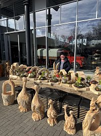 Ein Stand mit verschiedenen Skulpturen aus Holz, dahinter steht eine Frau und lächelt in die Kamera.