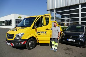 Ein Mitarbeiter des Autohaus Peter steht neben einem gelben ADAC-Servicefahrzeug mit ge&ouml;ffneter T&uuml;r. Im Hintergrund eine moderne Werkstatthalle mit Glasfronten und ein schwarzes Smart-Auto, links ein weiteres Geb&auml;ude.