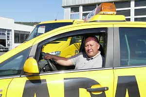 Ein Mitarbeiter des Autohaus Peter sitzt am Steuer eines gelben ADAC-Servicefahrzeugs mit gro&szlig;em ADAC-Schriftzug. Er schaut aus dem offenen Fenster und l&auml;chelt, im Hintergrund sind weitere gelbe Servicefahrzeuge sowie eine Werkstatthalle mit gro&szlig;en Fenstern zu sehen.