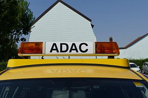 Nahaufnahme eines gelben Fahrzeugs mit ADAC-Schriftzug auf dem Dachbalken mit orangefarbenen Warnleuchten. Im Hintergrund sind H&auml;user mit hellen Fassaden sowie B&auml;ume zu sehen.
